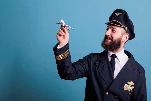 Улыбающийся пилот в форме, держащий модель самолета, авиатор академии, играющий с самолетом. Член экипажа самолета смотрит на коммерческую реактивную игрушку, боковой вид студии средний снимок - Фото, изображение