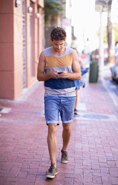 Επικοινωνώ εν κινήσει. Πλήρης εικόνα ενός νεαρού να περπατάει στο δρόμο της πόλης στέλνοντας μηνύματα στο κινητό του. - Φωτογραφία, εικόνα