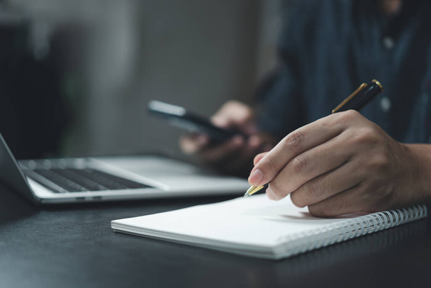 Ο άνθρωπος χέρι γράφει σε ένα σημειωματάριο με ένα στυλό και ένα έξυπνο τηλέφωνο στο γραφείο. - Φωτογραφία, εικόνα