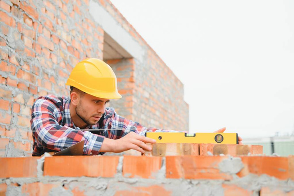 Using bricks. Young construction worker in uniform is busy at the unfinished building. - Photo, image