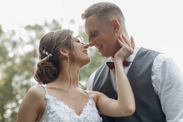 Side view of happy smiling wedding couple standing outside near trees. Young good-looking woman bride touching neck of young beautiful man groom, meeting eyes, posing. Relationship, love, wedding. - Fotografie, Obrázek