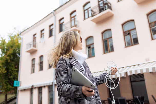бізнес-леді в місті на вулиці з ноутбуком в руках шукає місце, де вона може заряджати свій ноутбук зарядним пристроєм в руках. - Фото, зображення