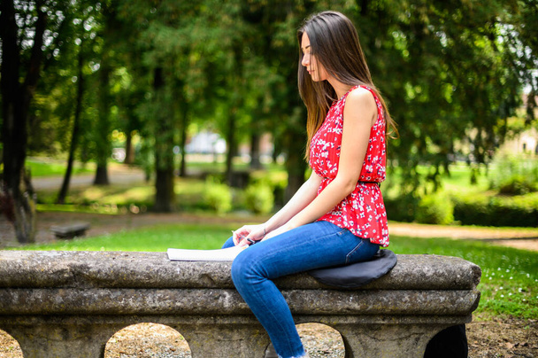 Beautiful female college student reading a book on a bench in a park - Фото, изображение