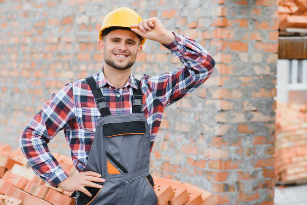 Construction worker man in work clothes and a construction helmet. Portrait of positive male builder in hardhat working at construction site - Zdjęcie, obraz