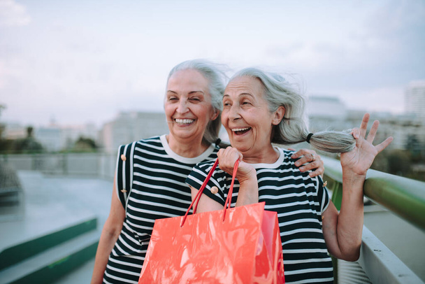 Happy senior twins in same clothes walking in a city, returning from shopping. - Фото, изображение