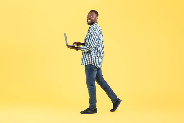 Musta mies, joka käyttää kannettavaa tietokonetta hymyillen kameralle Posing Walking In Studio Over Yellow Background. Freelance and Internet Technology konsepti. Täyspitkä laukaus - Valokuva, kuva