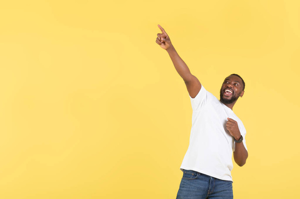 Kijk daar. Opgewonden Afro-Amerikaanse man wijzend vinger omhoog poseren in de buurt van lege ruimte voor tekst reclame grote aanbod staande over gele achtergrond. Studio neergeschoten. - Foto, afbeelding