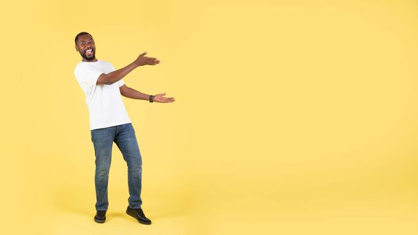見ろ!。興奮アフリカ系アメリカ人男性両方の手でテキストのための空白スペースを表示カメラに笑みを浮かべて黄色の背景に立ってポーズ.人の広告の大きい提供。パノラマ - 写真・画像