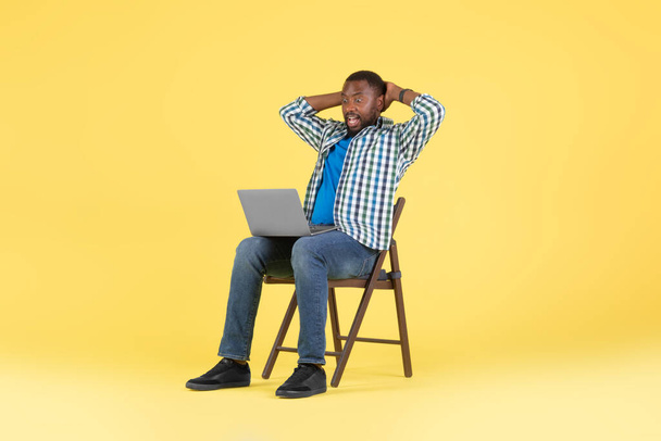 ラップトップコンピュータを見て感情的な黒人男性読書素晴らしいオンラインオファー黄色の背景、スタジオショットの上に椅子に座って作業を提供しています。素晴らしいウェブサイト、インターネット技術の概念 - 写真・画像