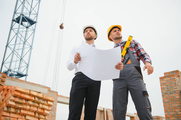 engineer and builder in hard hats discussing blueprint on construction site - Zdjęcie, obraz
