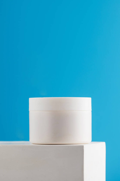 Burla de un frasco blanco con crema nutritiva para la cara y el cuerpo sobre un pedestal sobre un fondo azul. Cuidado de la piel. Copiar espacio - Foto, Imagen