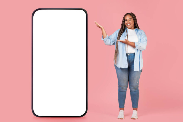 Happy Black Lady Shooting Huge с пустым экраном, рекламирующим новое мобильное приложение Smiling to Camera, стоящее на розовом студийном фоне. Полная длина, макет - Фото, изображение