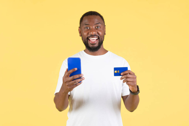 Hombre negro feliz usando la aplicación móvil de compras vía Smartphone y tarjeta de crédito sonriendo a la cámara que hace el pago en línea en fondo amarillo del estudio. Comercio electrónico, concepto de aplicación bancaria - Foto, imagen