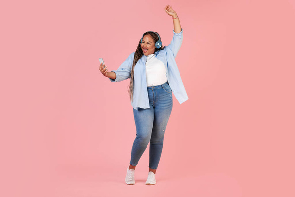 喜びのプランプアフリカ系アメリカ人女性は、ピンクの背景の上に置かれたヘッドフォンを身に着けているスマートフォンを介して音楽を聴いて踊る。音楽アプリケーションの概念。フルレングス、スタジオショット - 写真・画像