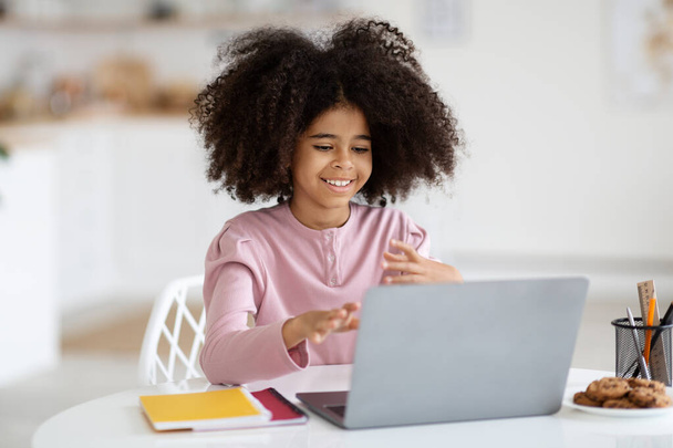 Nettes fröhliches afrikanisch-amerikanisches kleines Schulmädchen mit Laptop zu Hause, Schulkind bei den Hausaufgaben, Frühchen bei der Arbeit an Bildungsprojekten, Kopierraum. Heimschule, e-Education-Konzept - Foto, Bild