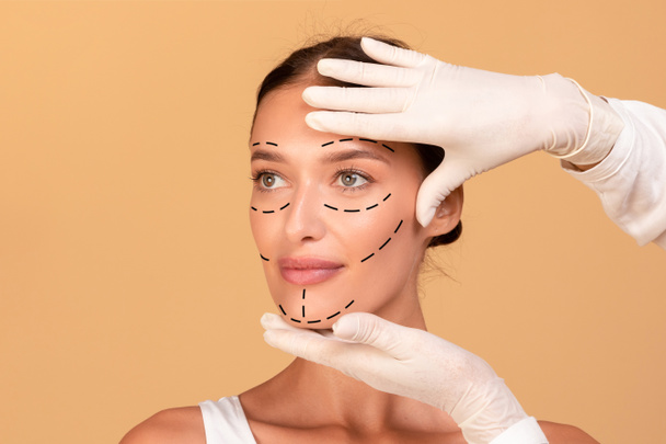 Frontális portré fiatal nő kap spa kezelés kozmetikus védőkesztyűben. Plasztikai sebész nyomot hagy a női arcon a bézs stúdió felett - Fotó, kép