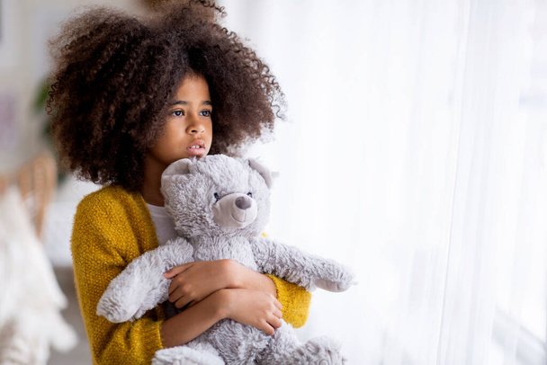 Closeup photo of sad african american preteen girl with bushy hair holding teddy bear and looking through the window, missing her friends while staying home during pandemic, copy space - Zdjęcie, obraz