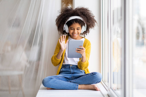 Happy african american child cute preteen girl with bushy hair sitting on windowsill at home, using wireless headphones and modern digital tablet, having online party with friends, copy space - Photo, Image