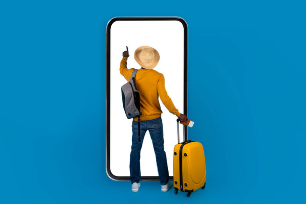 Takaisin näkymä musta mies turisti yllään kesähattu, kuljettaa reppu ja keltainen matkatavarat käyttäen valtava älypuhelin valkoinen tyhjä näyttö sininen tausta, varaus hotelli tai lippuja verkossa, mockup - Valokuva, kuva