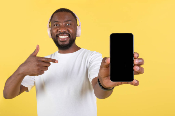 無線イヤフォンを装着した携帯電話のブランク画面を表示する幸せなアフリカ系アメリカ人男性スタジオで黄色の背景にある携帯電話で指を指す音楽アプリケーションを宣伝します。選択的フォーカス - 写真・画像