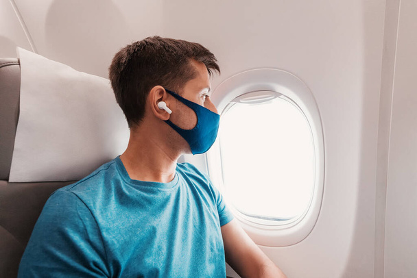Junger Mann reist mit dem Flugzeug, trägt während der Covid-19-Pandemie eine Gesichtsmaske, hört Musik mit Kopfhörern und blickt aus dem Fenster - Foto, Bild