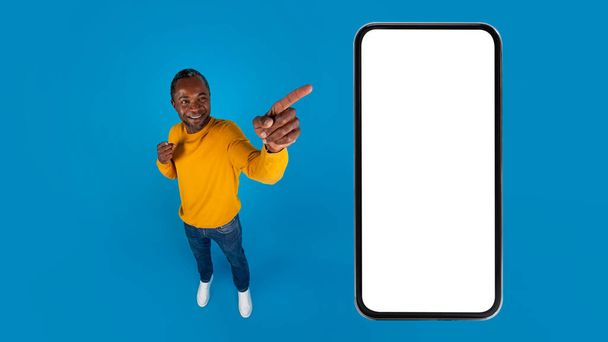 白いスクリーン、青いスタジオの背景、高い角度のビュー、コピースペースのパノラマ、広告のためのモックアップを持つ巨大なスマートフォンを指してカジュアルな服装で中年の黒人男性を笑顔 - 写真・画像