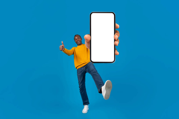 Cheerful mature black man in casual showing brand new cell phone with white blank screen and gesturing thumb up, lifting leg up, blue studio background, panorama with copy space, mockup, full length - Photo, Image