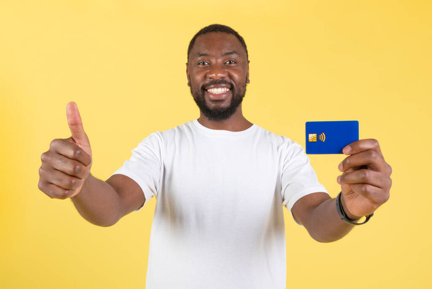 Ich mag meine Bank. Fröhlicher afroamerikanischer Typ, der mit Kreditkarte gestikulierende Daumen nach oben zeigt und lächelnd vor der Kamera über gelbem Studiohintergrund posiert. Männliche Zustimmung zu großem Finanzangebot - Foto, Bild