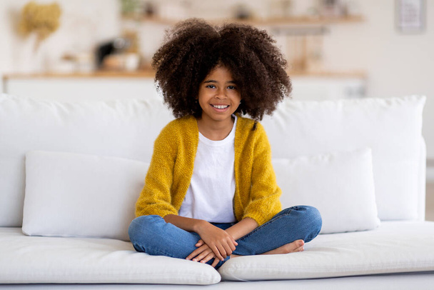 Wesoła, afrykańska dziewczyna siedząca na kanapie w domu, szczęśliwa, słodka, czarna dziewczynka z krzaczastymi włosami spoczywająca na białej kanapie, uśmiechnięta do kamery, przestrzeń do kopiowania. Koncepcja stylu życia dzieci - Zdjęcie, obraz