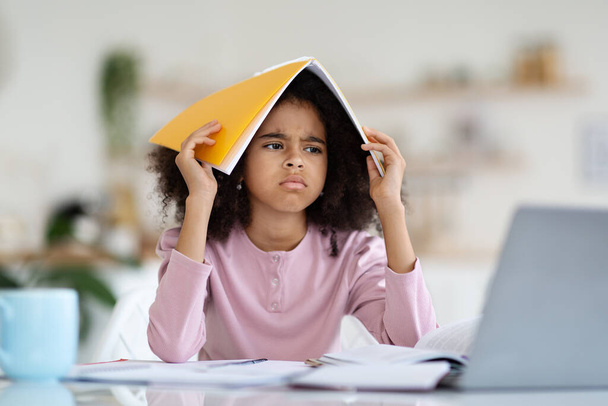 Angry preteen black girl with adorable bushy hair schooler doing homework at home, sitting at table, holding notepad on her head, looking at laptop screen, having difficulties, copy space - Photo, Image