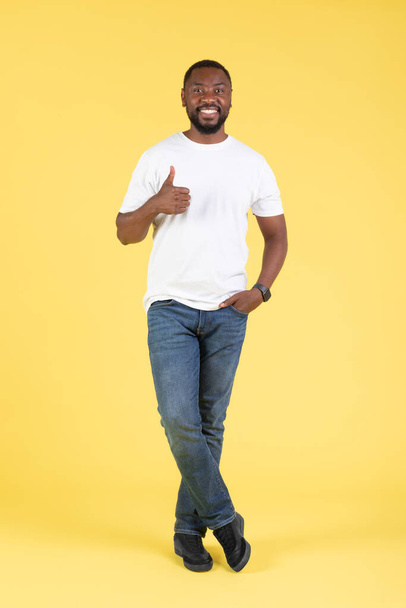Pozytywny czarny facet Gesturing kciuki w górę pozowanie uśmiech do kamery stojąc w studio na żółtym tle. Mężczyzna pokazuje się jak w aprobacie świetnej oferty. Strzał pionowy - Zdjęcie, obraz