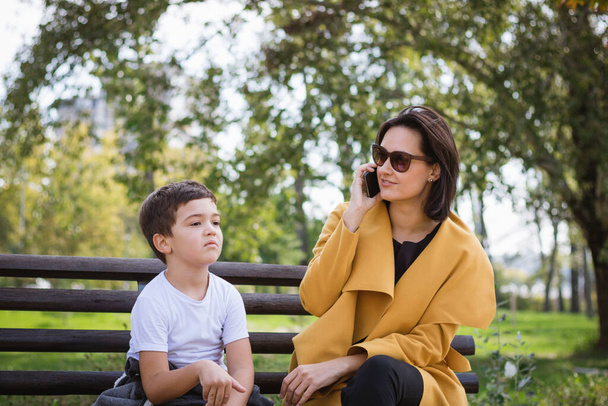 Маленький хлопчик почувається нудним, сидячи зі своєю матір'ю на лавці парку і чекаючи, поки вона закінчить телефонний дзвінок
.  - Фото, зображення