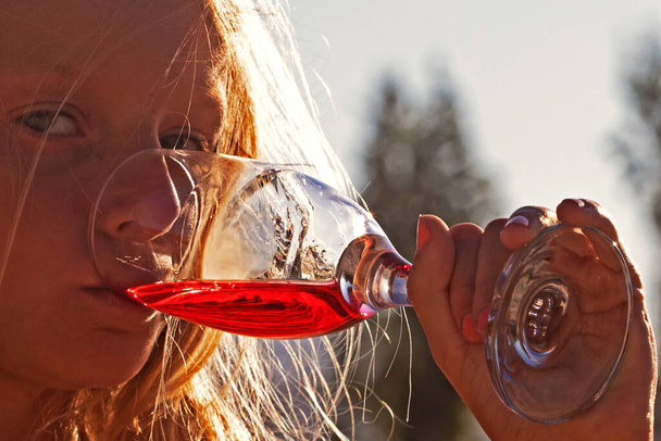 Egy kislány vörös italt iszik egy pohárból a nap ellen egy partin.. - Fotó, kép