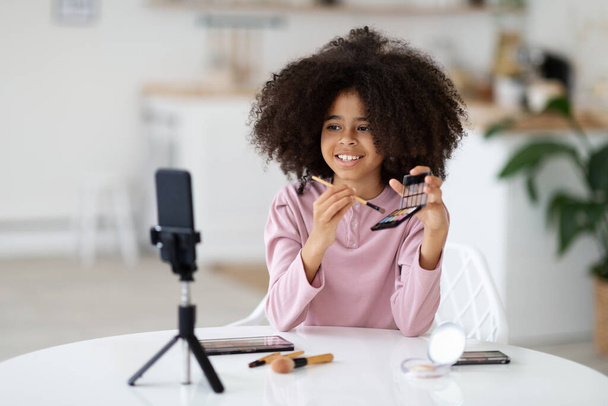 Beauty blog for kids concept. Pretty cute black preteen girl recording video for her vlog, using smartphone set on tripod, child sitting at table at home, showing fancy cosmetics for little girls - Photo, Image