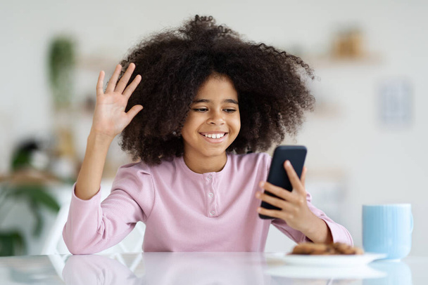 Cheerful african american girl with bushy hair having video call, sitting at desk, eating cookies, drinking juice or cocoa, holding mobile phone, waving at gadget screen and smiling, copy space - Fotó, kép