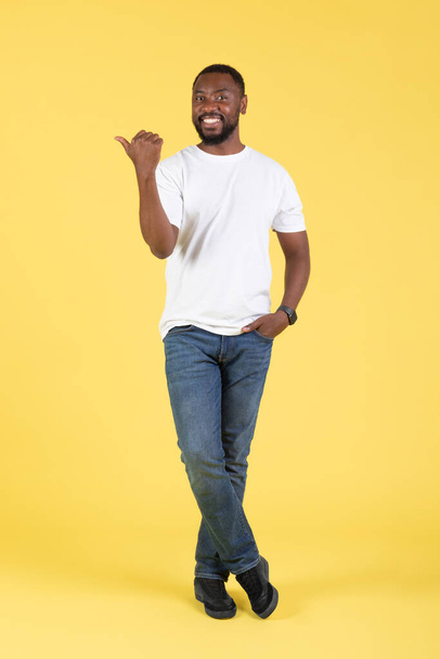 Kijk daar. Verticaal schot van vrolijke zwarte man wijzen duim vinger naast reclame aanbieden, glimlachend kijkend naar de camera staande over gele achtergrond. Studio neergeschoten, volledige lengte - Foto, afbeelding