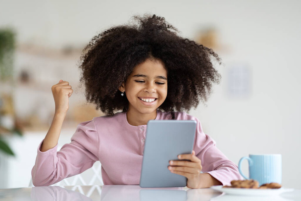 Happy African American preteen girl with cute bushy hair playing video game on digital tablet, κοιτάζοντας την οθόνη pad, σηκώνοντας το χέρι και χαμογελώντας, γιορτάζοντας τη νίκη, εσωτερικό σπίτι, αντίγραφο χώρο - Φωτογραφία, εικόνα