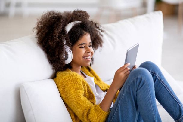 Smiling black child cute preteen girl sitting on sofa, using wireless headphones and modern digital tablet, watching cartoons on Internet, home interior, side view, copy space - Photo, Image