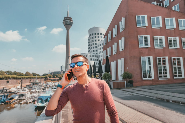 Hombre feliz hablando por teléfono inteligente de la famosa torre de televisión Dusseldorf desde el canal de Media Harbor en el distrito post industrial. Lugares de viaje y turismo - Foto, Imagen