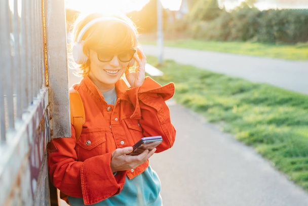 Smiling stylish woman in bright clothes and sunglasses wearing wireless headphones on her head and seting phone for listening music. Walk in city park on sunset. Hipster lifestyle. Selective focus. - 写真・画像