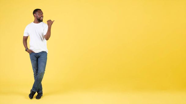 幸せなアフリカ系アメリカ人男性は、黄色の背景に立ってテキスト広告のためのフリースペースで親指を脇に向けて,スタジオショット.男の広告何か。コンセプトがある。パノラマ - 写真・画像