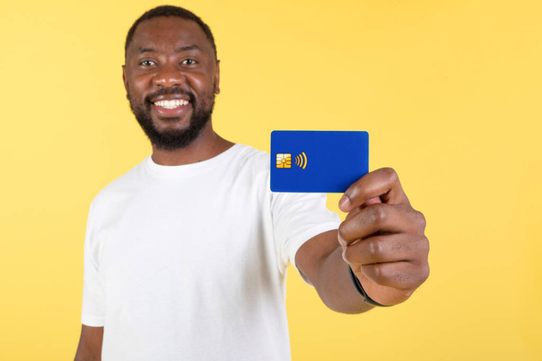 Smiling Black Man Showing His Credit Card To Camera Advertising Great Bank Offer Posing Standing On Yellow Background, Studio Shot. Payment And Financial Safety Concept. Selective Focus - Foto, immagini