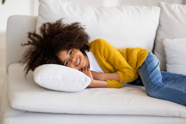 Niño afroamericano relajado acostado en el sofá en casa, chica negra linda feliz con el pelo tupido reclinado en un sofá blanco y sonriendo, descansando, espacio de copia. concepto de estilo de vida infantil - Foto, Imagen