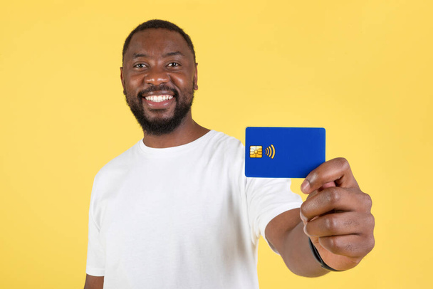 Allegro ragazzo afroamericano che mostra carta di credito Raccomandare offerta bancaria in piedi in posa in studio su sfondo giallo. Concetto di investimento in denaro e finanza - Foto, immagini