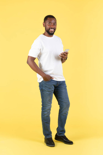 Allegro uomo nero utilizzando Smartphone in piedi che comunica SMS online su sfondo giallo Studio. Ragazzo pubblicità grande applicazione mobile per il telefono. Colpo verticale - Foto, immagini