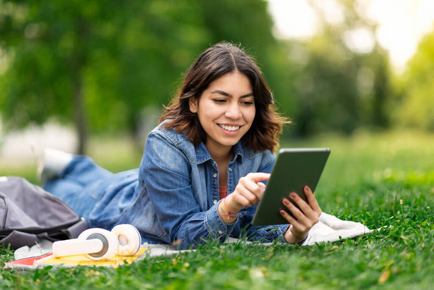 Sorrindo jovem árabe mulher que navega nas mídias sociais em tablet digital enquanto relaxa ao ar livre no parque da cidade, feliz estudante do Oriente Médio deitado no gramado e usando gadget moderno, espaço cópia - Foto, Imagem
