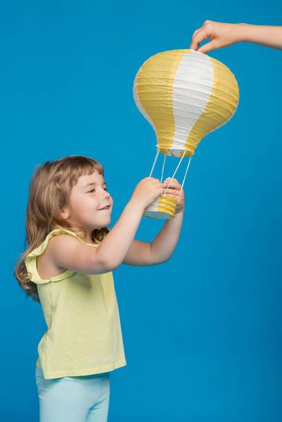little girl touches yellow balloon, hands raised, looks and smiles, concept of adventure and fantasy, on blue background - Photo, Image