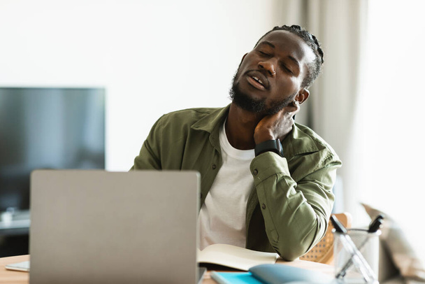 Zmęczony czarny człowiek cierpiący na skurcze szyi podczas pracy na laptopie w biurze, siedzi przy biurku i masuje mięśnie. Afrykański Amerykanin ma skurcz szyi spowodowany niewygodną pozycją - Zdjęcie, obraz