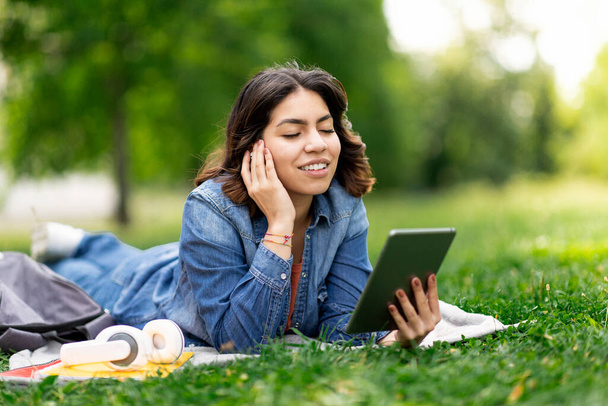Beautiful Young Arab Woman In Earphones Listening Music On Digital Tablet Outdoors, Smiling Middle Eastern Female Using Modern Gadget, Enjoying Favorite Playlist While Lying On Lawn In City Park - 写真・画像