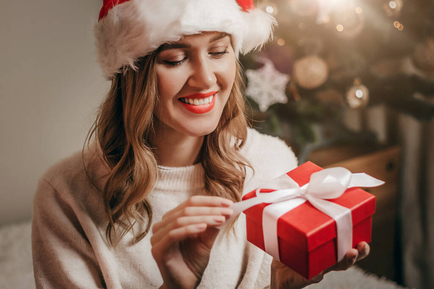 Boldog nő egy újévi jelmezben mosolyog és kipakol egy ajándékdobozt az újévi dekorációk hátterére és egy karácsonyfát koszorúkkal. - Fotó, kép